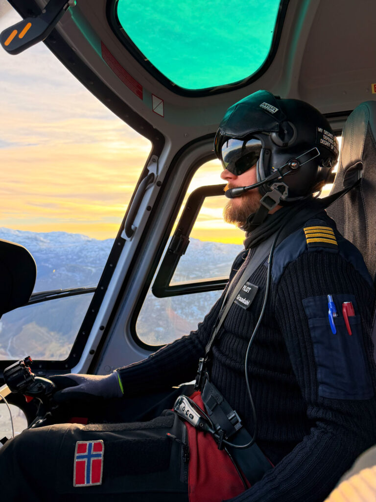 Bilde av en helikopterpilot som sitter med hjelm og briller og kjører i solnedgangen